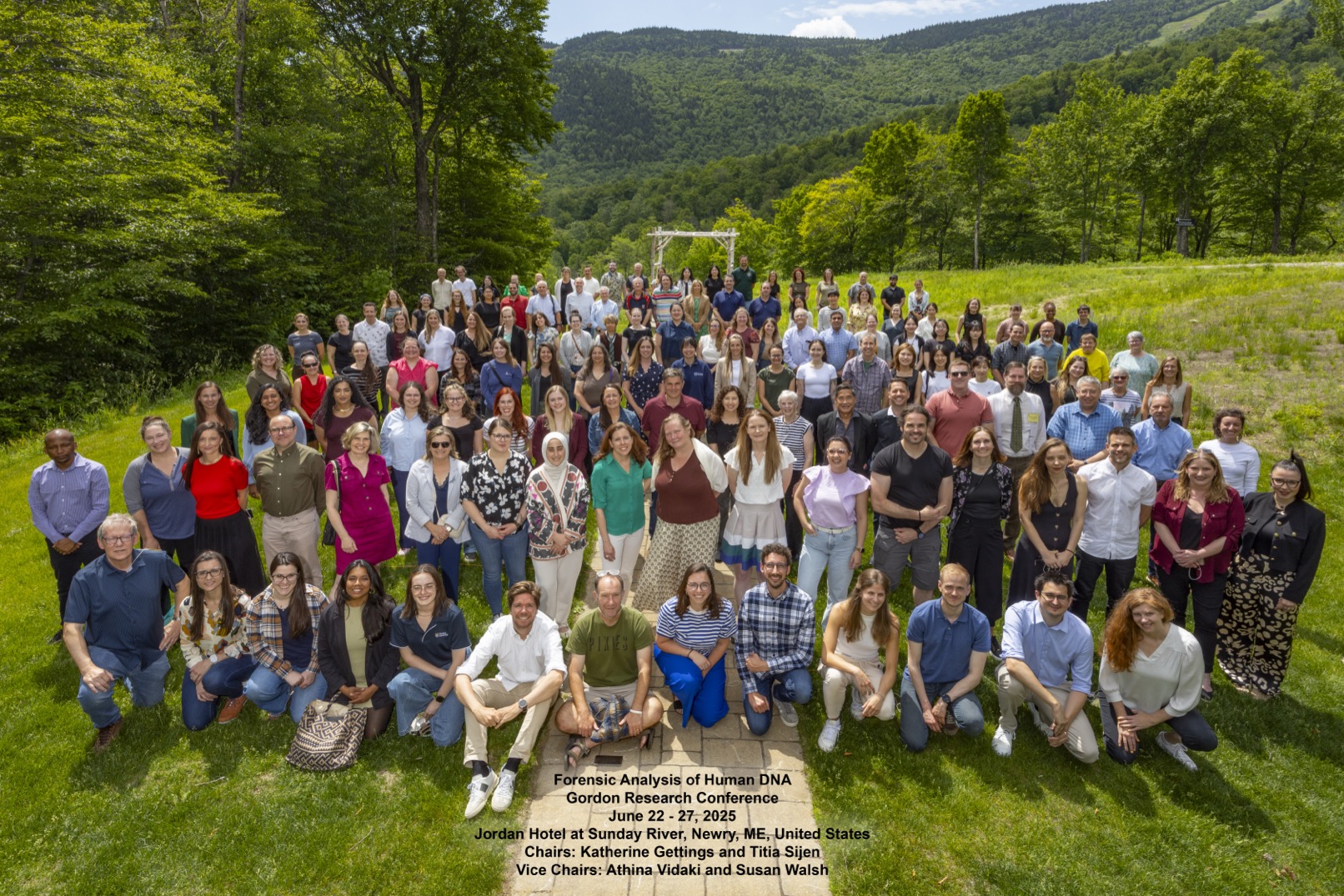 Grupa ludzi siedzących i stojących na trawie, w tle drzewa i góry