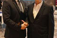 Dwóch mężczyzn w garniturach stoi obok siebie w sali konferencyjnej i uśmiecha się do zdjęcia; w tle widoczny ekran prezentacyjny