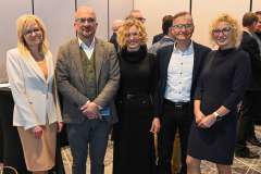 Trzy kobiety i dwóch mężczyzn stoi obok siebie w sali konferencyjnej i pozuje do zdjęcia; w tle widoczni są inni uczestnicy wydarzenia.