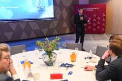 Mężczyzna w garniturze stoi przy ekranie prezentacyjnym i przemawia do uczestników siedzących przy stole