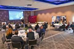 Mężczyzna w garniturze stoi przy ekranie prezentacyjnym i przemawia do uczestników siedzących przy stołach