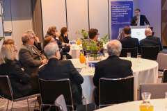 Mężczyzna w garniturze stoi przy ekranie prezentacyjnym i przemawia do uczestników siedzących przy stołach