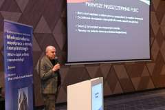 Mężczyzna stoi przy mównicy i przemawia do uczestników konferencji; za nim na ekranie wyświetlona jest prezentacja 