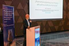 Mężczyzna stoi przy mównicy i przemawia do uczestników konferencji; za nim na ekranie wyświetlona jest prezentacja 