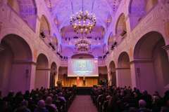 Widok wnętrza dużej sali z publicznością siedzącą na widowni i sceną na końcu, oświetlonej fioletowo. Na scenie stoją dwie osoby