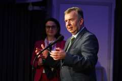 Mężczyzna w garniturze stoi przy mikrofonie podczas uroczystego wystąpienia, z tyłu stoi kobieta i trzyma teczkę z dokumentami