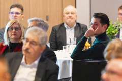 Uczestnicy konferencji siedzą przy okrągłych stolikach; na pierwszym planie młody mężczyzna w zielonym swetrze słucha prelegenta, 