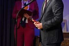 Mężczyzna w garniturze stoi przy mikrofonie podczas uroczystego wystąpienia, z tyłu stoi kobieta i trzyma teczkę z dokumentami