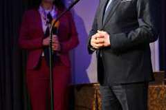 Mężczyzna w garniturze stoi przy mikrofonie podczas uroczystego wystąpienia, z tyłu stoi kobieta i trzyma teczkę z dokumentami