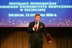 Mężczyzna w garniturze stoi na scenie z mikrofonem i teczką, przemawiając podczas Spotkania Noworocznego