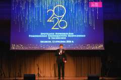 Mężczyzna w garniturze stoi na scenie z mikrofonem i teczką, przemawiając podczas Spotkania Noworocznego