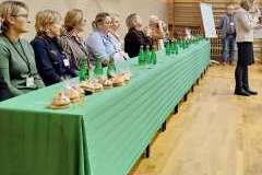 jury zasiadające za stołem, przed stołem stoi kobieta i mówi przez mikrofon