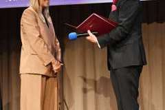 Mężczyzna trzyma mikrofon i czerwoną teczkę, stojąc naprzeciw kobiety w jasnym garniturze na scenie podczas uroczystości