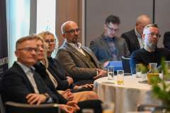 Grupa uczestników konferencji siedzi przy okrągłym stole i słucha wystąpienia; na stole stoją szklanki z napojami i materiały informacyjne