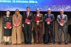 Grupa osób stoi na scenie z bordowymi teczkami, pozując do zdjęcia podczas uroczystego wydarzenia