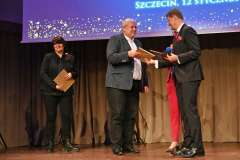 Jeden mężczyzna podaje do ręki wyróżnienie drugiemu mężczyźnie na scenie, jeden z nich trzyma dokument, podczas uroczystej ceremonii