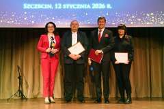 Grupa osób stoi na scenie z bordowymi teczkami, pozując do zdjęcia podczas uroczystego wydarzenia