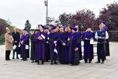 Grupa osób w fioletowych togach i biretach akademickich stoi na placu na zewnątrz budynku. Część z nich trzyma dokumenty