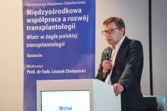 Mężczyzna przemawia przy mównicy podczas konferencji; obok widoczny baner z tytułem wydarzenia dotyczącym współpracy w transplantologii