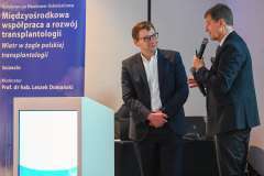 Dwóch mężczyzn w garniturach stoi przy mównicy podczas konferencji; jeden trzyma mikrofon, w tle widoczny baner wydarzenia 