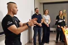 dwie kobiety i dwóch mężczyzn stojących przed ekranem monitora, jeden mężczyzna gestykuluje