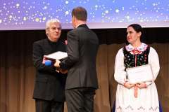 Dwóch mężczyzn podaje sobie ręce na scenie podczas uroczystej ceremonii, obok stoi kobieta w stroju ludowym z oprawionym wyróżnieniem w ręku