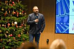 mężczyzna na scenie mówiący przez mikrofon, w tle ekran z prezentacją oraz choinka