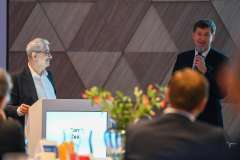 Dwóch mężczyzn stoi przy podium podczas konferencji; jeden przemawia do mikrofonu, drugi słucha. Na pierwszym planie widoczni są uczestnicy 