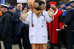 Osoba przebrana za maskotkę pumy w białym fartuchu stoi na placu wśród uczestników wydarzenia akademickiego. Wokół widoczne są osoby w togach 