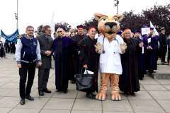 Osoba przebrana za maskotkę pumy w białym fartuchu stoi na placu wśród uczestników wydarzenia akademickiego. Wokół widoczne są osoby w togach 