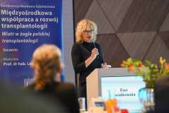 Kobieta przemawia przy mównicy podczas konferencji; obok widoczny baner z tytułem wydarzenia dotyczącym współpracy w transplantologii