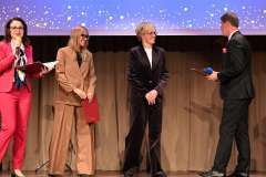 Cztery osoby stoją na scenie podczas uroczystości; mężczyzna w garniturze zwraca się do trzech kobiet, z których dwie trzymają teczkę
