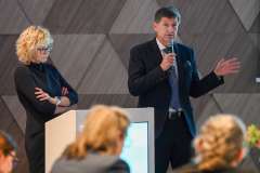 Mężczyzna z mikrofonem przemawia podczas konferencji; obok niego przy podium stoi kobieta, a na pierwszym planie widoczni są słuchacze