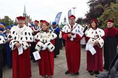 Grupa władz uczelni w czerwonych togach z białymi gronostajowymi pelerynami i czerwonych biretach stoi na placu podczas uroczystości akademickiej