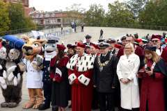 Grupa przedstawicieli władz uczelni w czerwonych togach z gronostajowymi pelerynami, osoby w mundurach oraz uczelniane maskotki stoją razem na placu 
