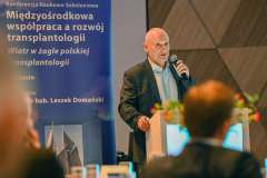 Mężczyzna z mikrofonem przemawia przy podium podczas konferencji; w tle widoczny jest baner z tytułem wydarzenia dotyczącym transplantologii