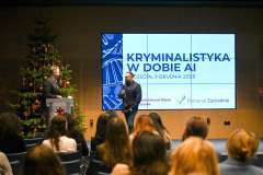 uczestnicy konferencji słuchają dwóch mężczyzn stojących i rozmawiających na scenie, w tle ekran wyświetlający prezentację