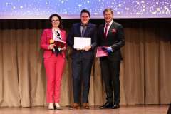 Trzy osoby stoją na scenie z wyróżnieniami w ramkach, pozując do zdjęcia podczas uroczystego wydarzenia