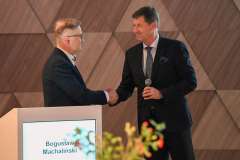 Dwóch mężczyzn w garniturach podaje sobie ręce przy podium konferencyjnym. Jeden z nich trzyma mikrofon