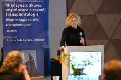 Kobieta z mikrofonem przemawia przy podium podczas konferencji; w tle widoczny jest baner z tytułem wydarzenia dotyczącym transplantologii