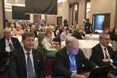 Uczestnicy konferencji siedzą przy okrągłych stołach w sali, słuchają i pracują na laptopach