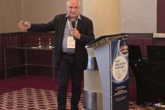 Mężczyzna przemawia przy mównicy podczas konferencji, trzyma mikrofon i gestem wskazuje w bok