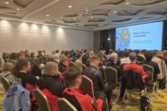 uczestnicy konferencji siedzący na sali konferencyjnej. Na ścianie ekran wyświetlający prezentację