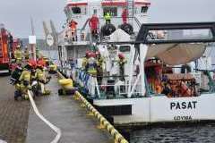 ratownicy straży pożarnej na statku, dwóch kuca przed statkiem trzymając wąż gaśniczy