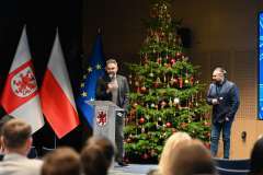 uczestnicy konferencji słuchają dwóch mężczyzn stojących i rozmawiających na scenie, w tle ekran wyświetlający prezentację