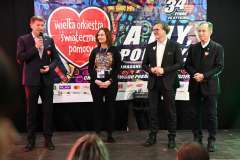 Cztery osoby stoją na scenie przed banerem Wielkiej Orkiestry Świątecznej Pomocy, mężczyzna przemawia do mikrofonu.