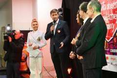 Trzech mężczyzn i dwie kobiety stoją na scenie Wośp, jeden mężczyzna mówi przez mikrofon. Widać dziennikarza z kamerą