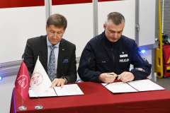 Dwóch mężczyzn podpisuje dokumenty przy stole, jeden w garniturze, drugi w mundurze policji.