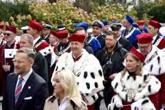 Władze uczelni w czerwonych togach z gronostajowymi pelerynami stoją wśród uczestników plenerowej uroczystości; w tle pozostali goście i zieleń