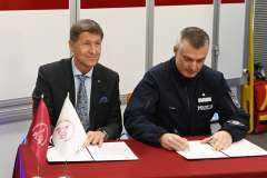 Dwóch mężczyzn podpisuje dokumenty przy stole, jeden w garniturze, drugi w mundurze policji.